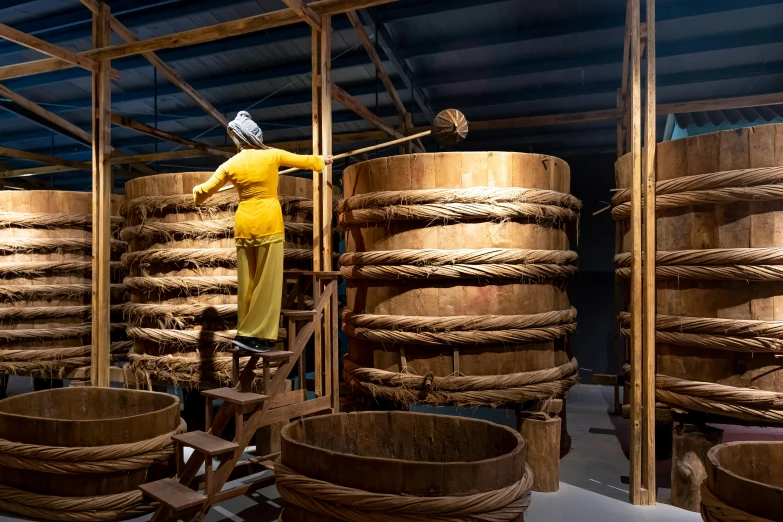 a man standing on a ladder in a room filled with wooden barrels, inspired by Li Di, process art, with yellow cloths, mill, hou china, a monumental