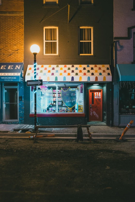 a bench sitting on the side of a street next to a building, by Drew Tucker, pexels contest winner, graffiti, convenience store, lights are on in the windows, colorful house, small library