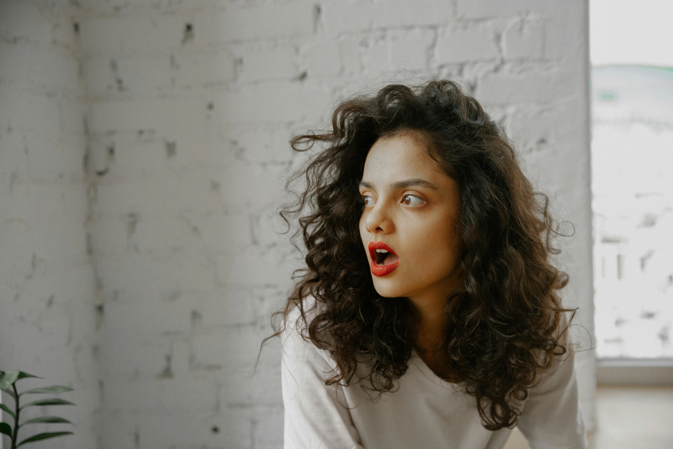 a woman with a surprised look on her face, by Emma Andijewska, trending on pexels, happening, long dark curly hair, bright red lipstick, dressed in a white t shirt, in an action pose