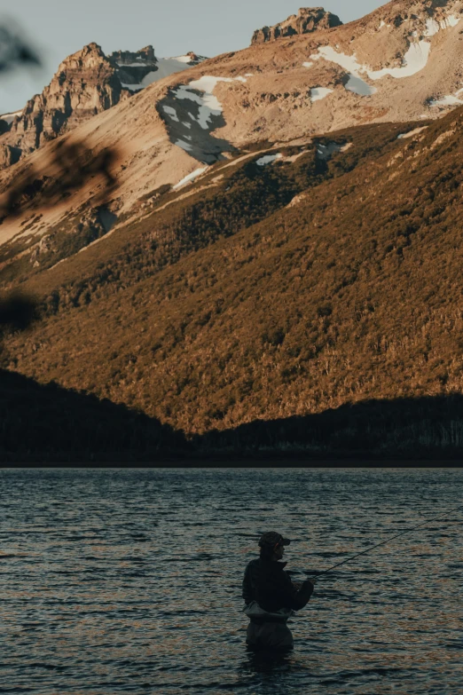 a person in a body of water with mountains in the background, a picture, late afternoon sun, patagonian, fishing, portrait featured on unsplash