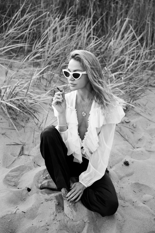 a woman sitting on top of a sandy beach, a black and white photo, inspired by Emma Andijewska, smoking a cigarrette🚬, with sunglass, beautiful young ornella muti, bone jewelry