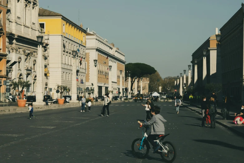 a man riding a bike down a street next to tall buildings, by Alessandro Allori, pexels contest winner, neoclassicism, kids playing, colosseo, square, yellow