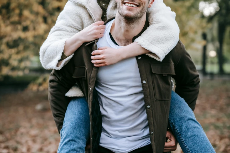 a man carrying a woman on his back in a park, trending on pexels, an aviator jacket and jorts, wearing hay coat, lachlan bailey, smiling man