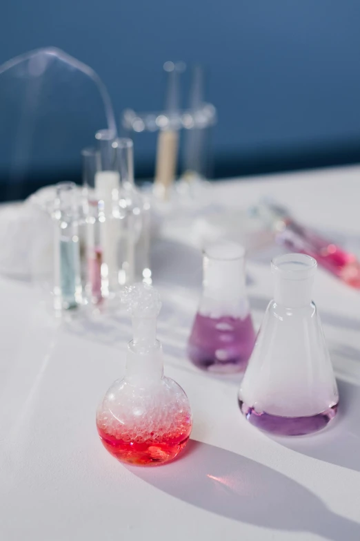 a group of flasks sitting on top of a white table, experimenting in her science lab, violet and pink palette, blue and white and red mist, with a lab coat