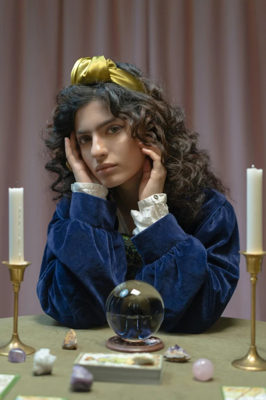a woman sitting at a table with a crystal ball, an album cover, inspired by Louis Le Nain, trending on pexels, renaissance, portrait androgynous girl, curls, beautiful jewish woman, gold and indigo