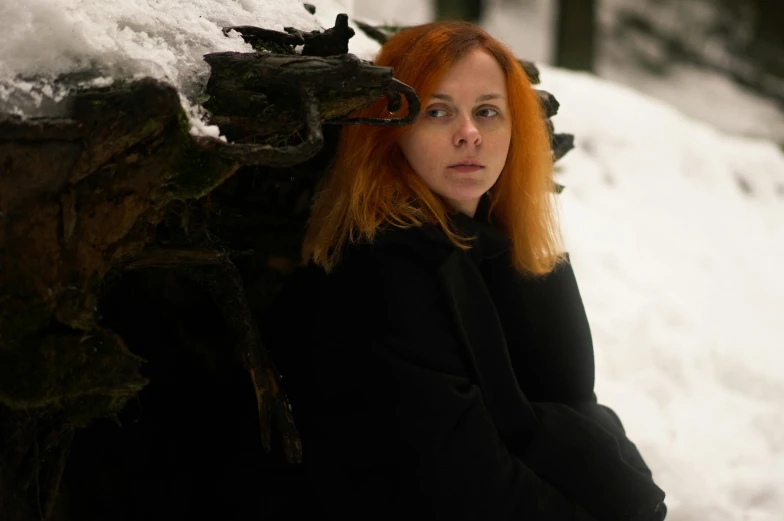 a woman leaning against a tree trunk in the snow, an album cover, inspired by Nína Tryggvadóttir, pexels contest winner, realism, orange hair, wearing black coat, ((portrait)), movie still 8 k