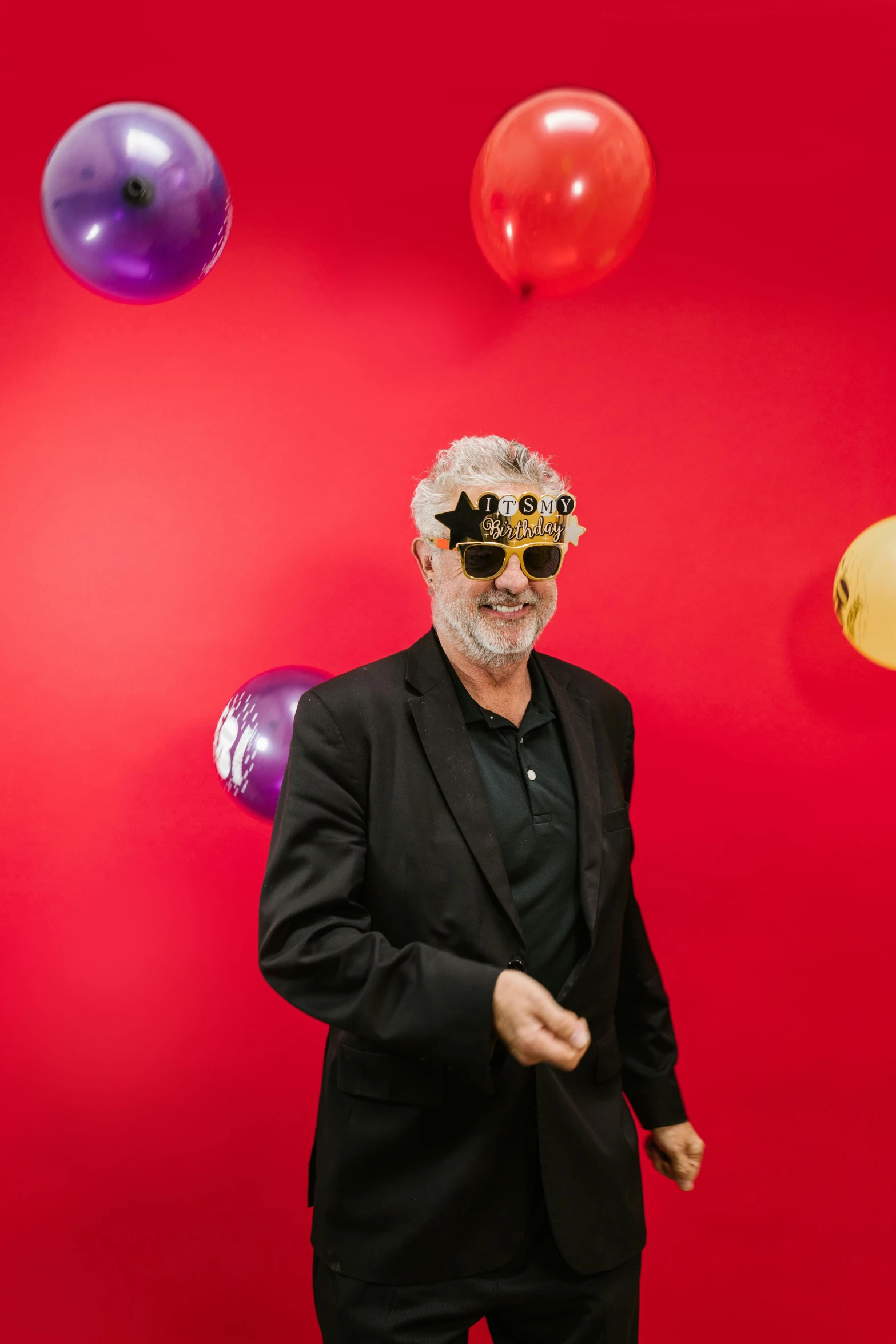 a man in a suit and sunglasses standing in front of balloons, grey beard, lighthearted celebration, mask off, boke