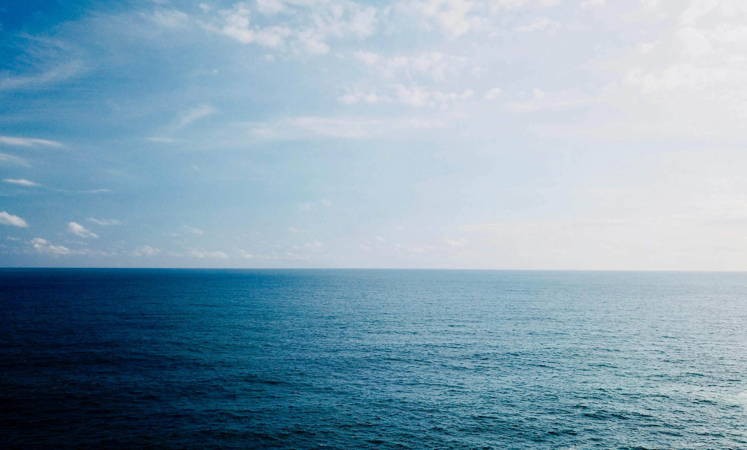 a large body of water next to a beach, inspired by Elsa Bleda, unsplash, minimalism, deep blue sea color, ignant, partly cloudy sky, rinko kawauchi