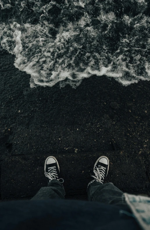 a person standing in front of a body of water, by Jacob Toorenvliet, unsplash contest winner, sneakers, looking down from above, black sand, towering waves