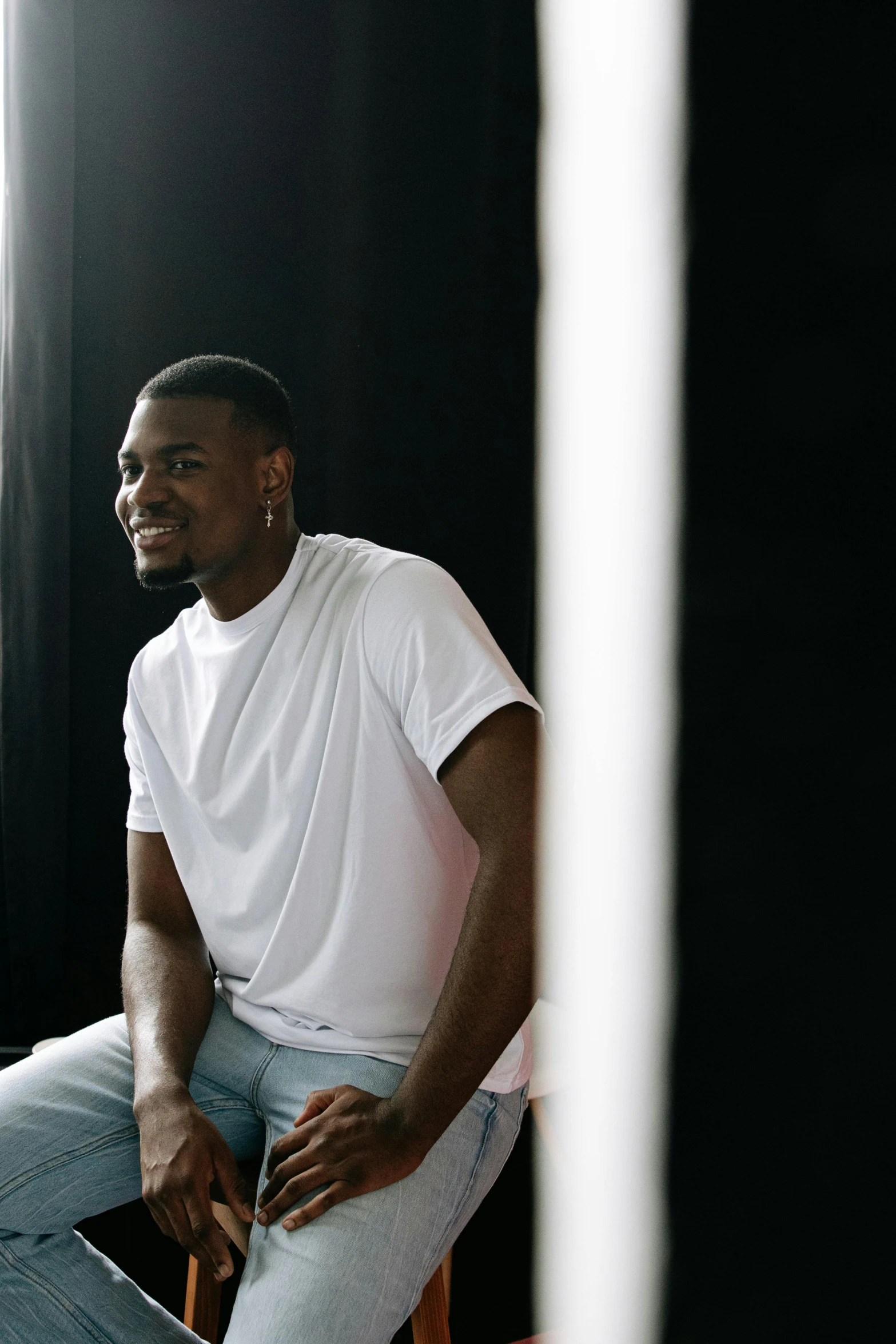 a man sitting on a chair in front of a window, a picture, inspired by Paul Georges, trending on unsplash, dressed in a white t shirt, jaylen brown, his smile threw shadows, studio spotlight