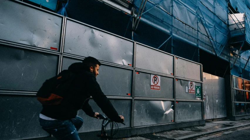 a man riding a bike down a street next to a tall building, pexels contest winner, graffiti, metal panels, worksafe. instagram photo, under construction, background image