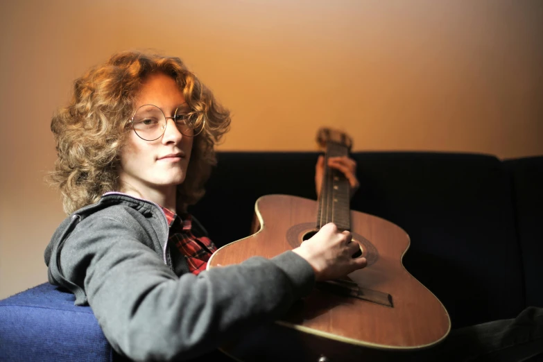 a woman sitting on a couch holding a guitar, inspired by Maxwell Gordon Lightfoot, profile image, androgynous person, looking towards camera, nerdy music teacher with phd