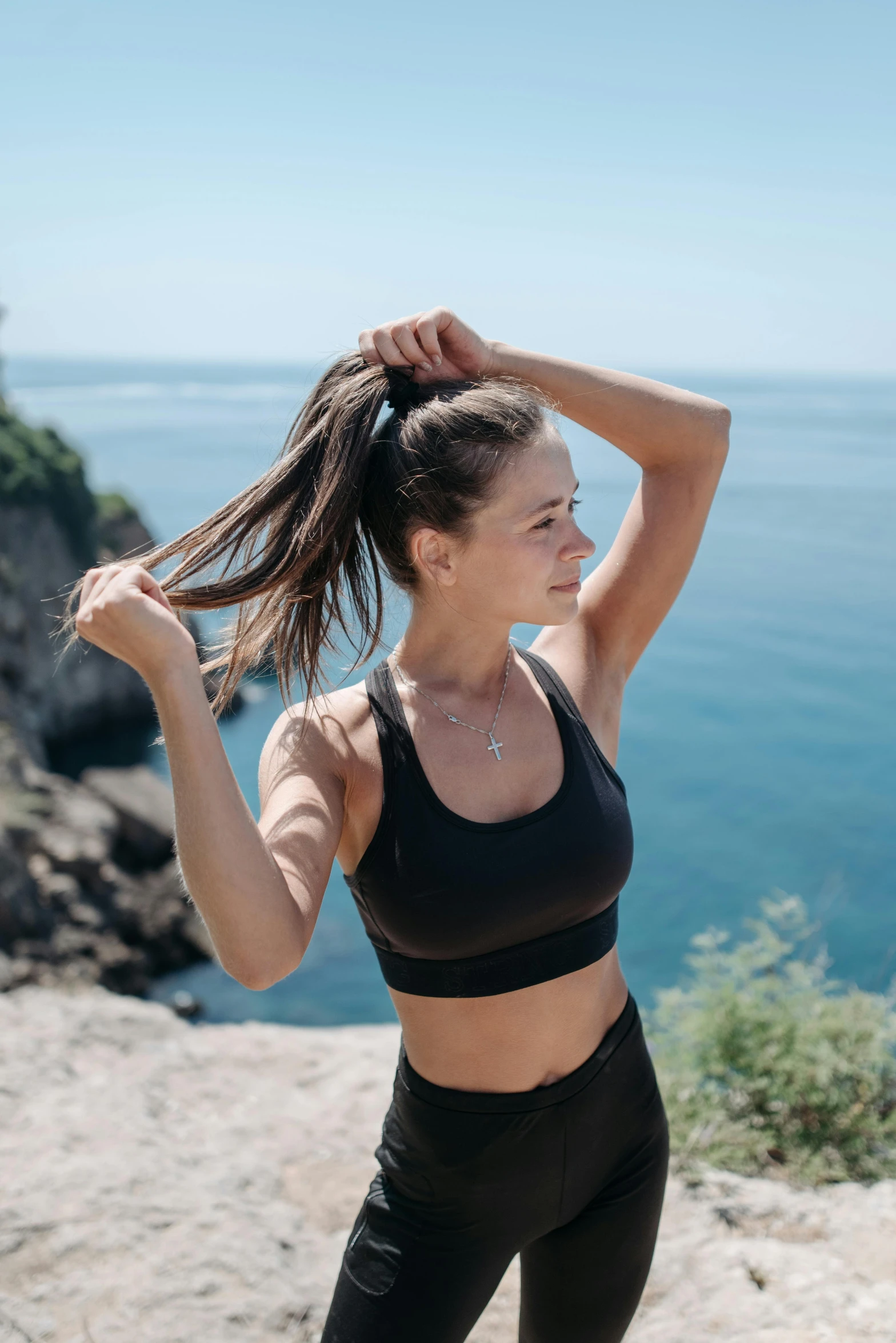 a woman standing on top of a cliff next to the ocean, sports bra, dynamic hair movement, her hair is tied above her head, profile image