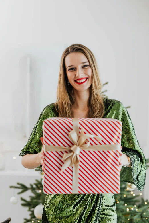 a woman holding a christmas present in front of a christmas tree, by Julia Pishtar, happening, cheeky smile with red lips, detailed product image, striped, helpful