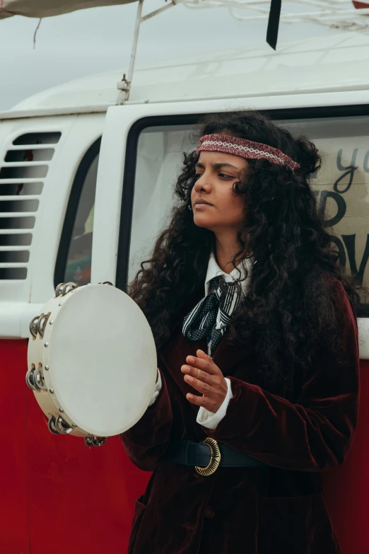 a woman standing next to a red and white van, an album cover, pexels contest winner, renaissance, playing drums, long dark curly hair, wearing a headband, caravan