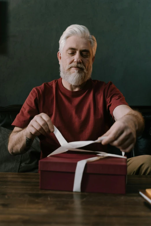 a man sitting at a table cutting a piece of paper, holding gift, very long silver beard, promo image, maroon