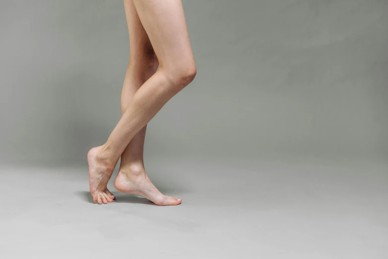 a woman standing in front of a gray background, inspired by Victorine Foot, pexels, arabesque, bare leg, thin young male, pale, engineered