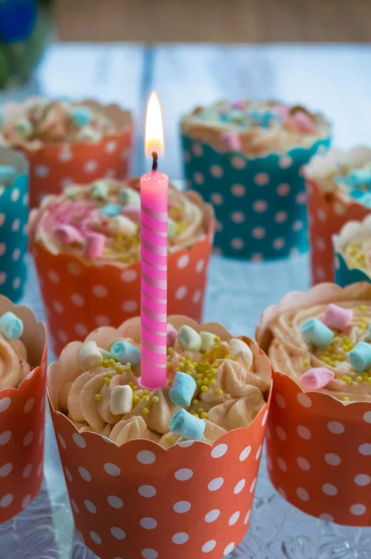 a table topped with cupcakes and a lit candle, pink and teal and orange, polka dot, medium close shot, square