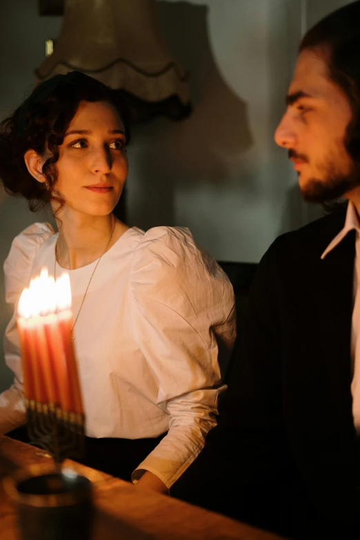 a man and a woman sitting at a table with a lit candle, a portrait, inspired by Ivan Kramskoi, pexels, beautiful jewish woman, production photo, looking off to the side, holiday season