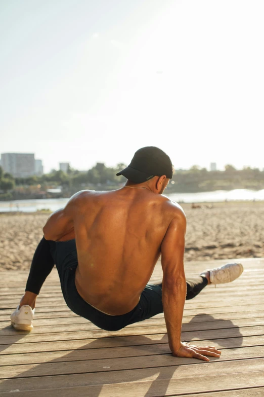 a man sitting on top of a wooden dock next to a body of water, dynamic stretching, fleshy musculature, at a beach, city views