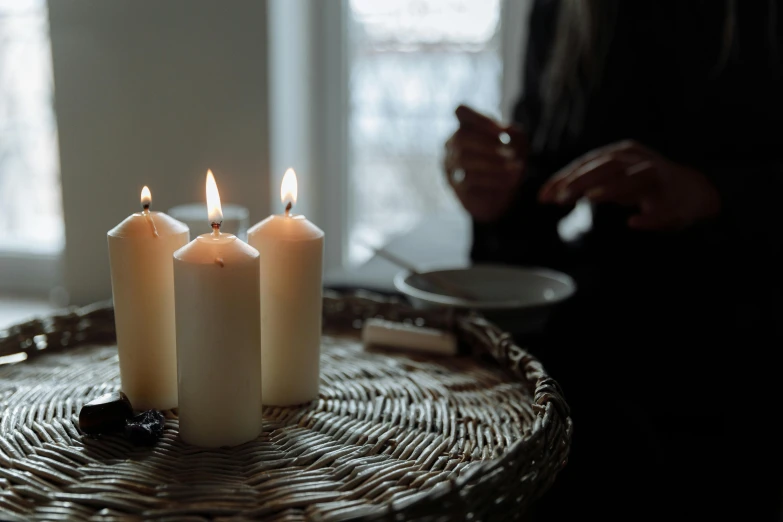 three white candles sitting on top of a wicker table, inspired by Elsa Bleda, unsplash, robed figures sat around a table, soft light from the window, ready to eat, amanda lilleston