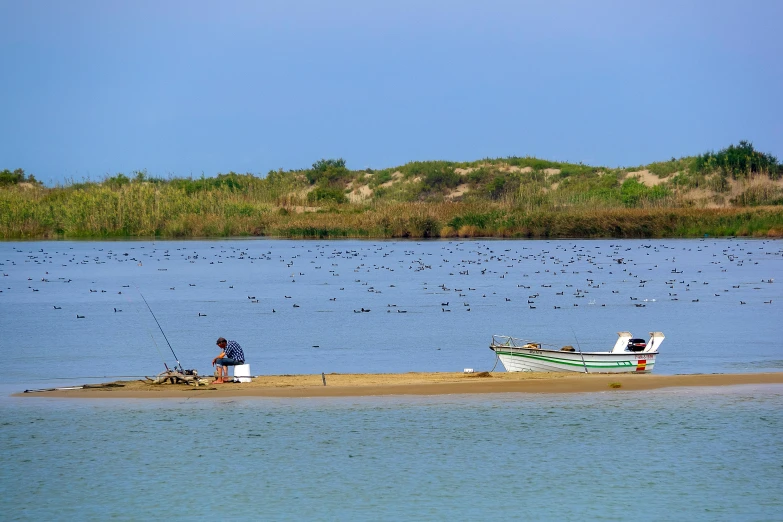 a man sitting on a beach next to a boat, by Peter Churcher, pixabay contest winner, hurufiyya, lagoon, fish flocks, farol da barra, viewed from a distance