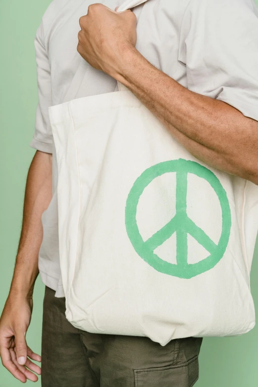 a man holding a bag with a peace sign on it, by Nina Hamnett, trending on pexels, private press, pastel green, cotton, irwin penn, plain background