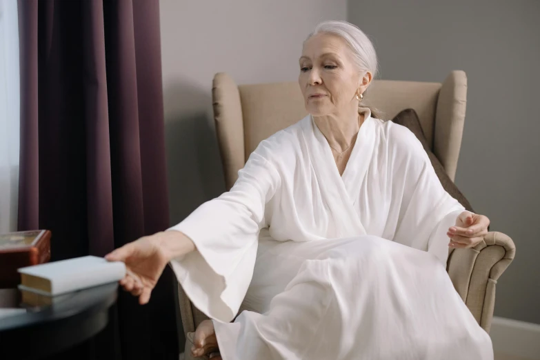 a woman in a white robe sitting in a chair, stretch, youtube thumbnail, elderly, wearing silver silk robe