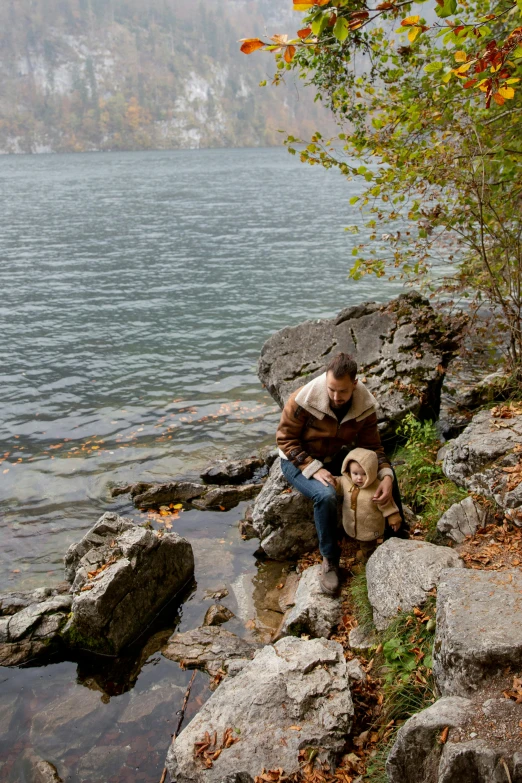 a man sitting on a rock next to a body of water, with dogs, during autumn, standing next to water, drinking
