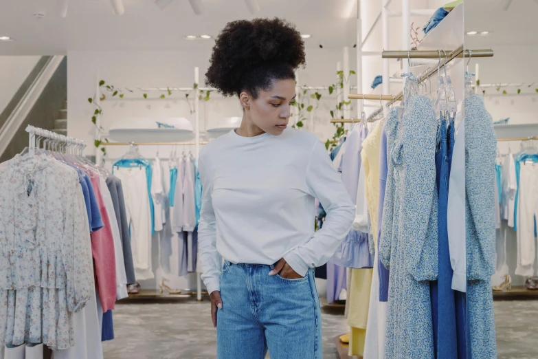 a woman standing in front of a rack of clothes, wearing a light blue shirt, ashteroth, white shirt and blue jeans, filmstill