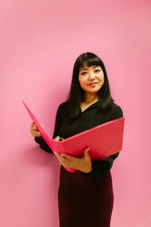 a woman standing in front of a pink wall holding a pink folder, inspired by Li Di, sui ishida with black hair, in an office, looking towards camera, brightly lit