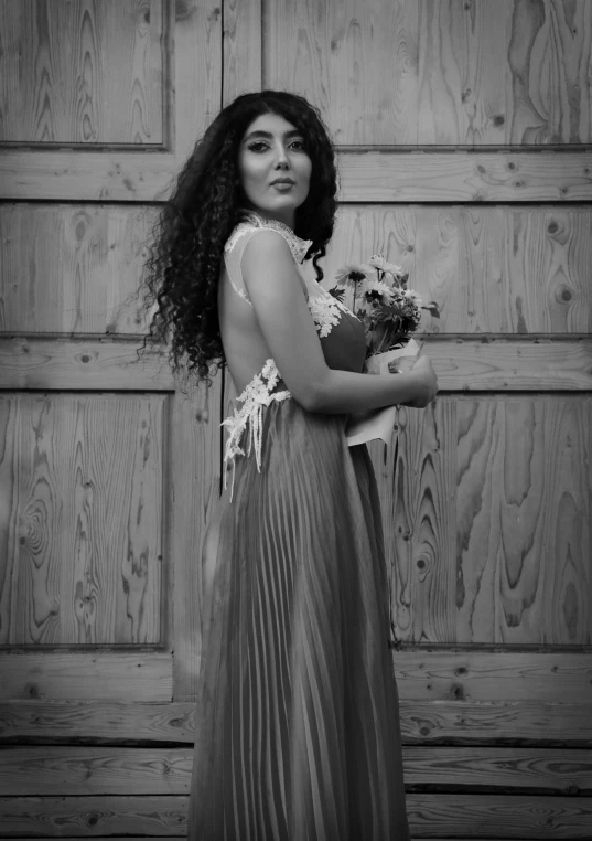 a woman standing in front of a wooden door, a black and white photo, inspired by irakli nadar, tumblr, romanticism, flowers!!!!, wearing silver dress, curly haired, monochrome:-2