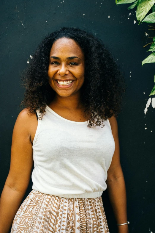 a woman standing in front of a black wall, by Olivia Peguero, dark short curly hair smiling, wearing white camisole, tan skin a tee shirt and shorts, long afro hair