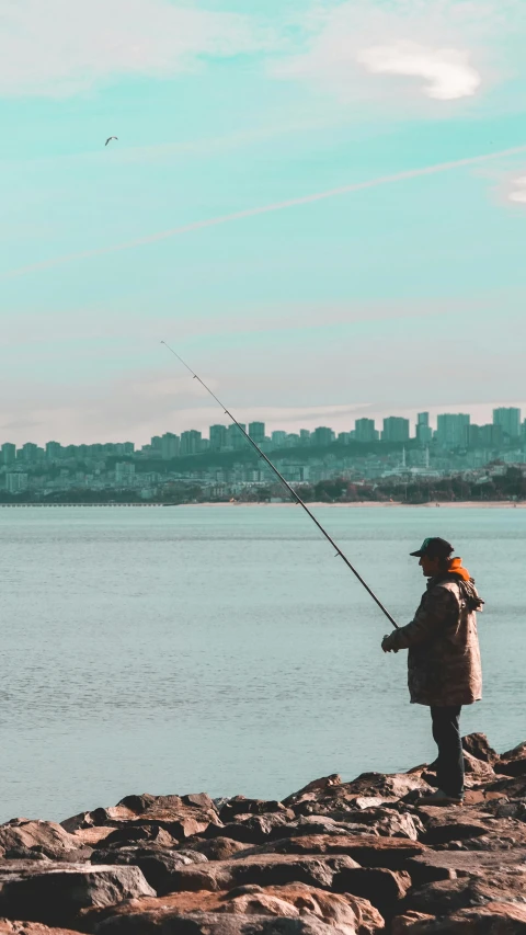 a man standing on top of a rock next to a body of water, fishing pole, cityscape, teal aesthetic, sports photo