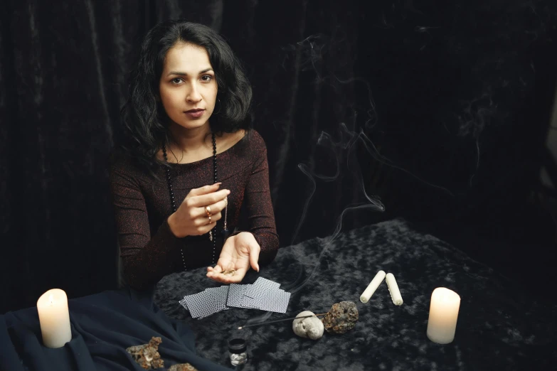 a woman sitting at a table with candles and playing cards, by Julia Pishtar, casting spells, on a gray background, woman in black robes, ameera al-taweel