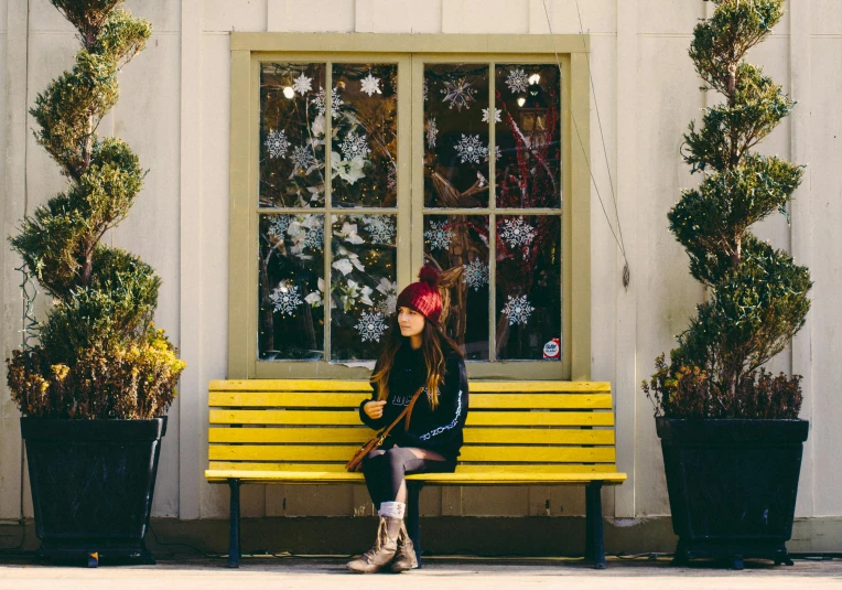 a woman sitting on a yellow bench in front of a building, by Julia Pishtar, pexels contest winner, wintertime, joanna gaines, holiday season, yellow cap