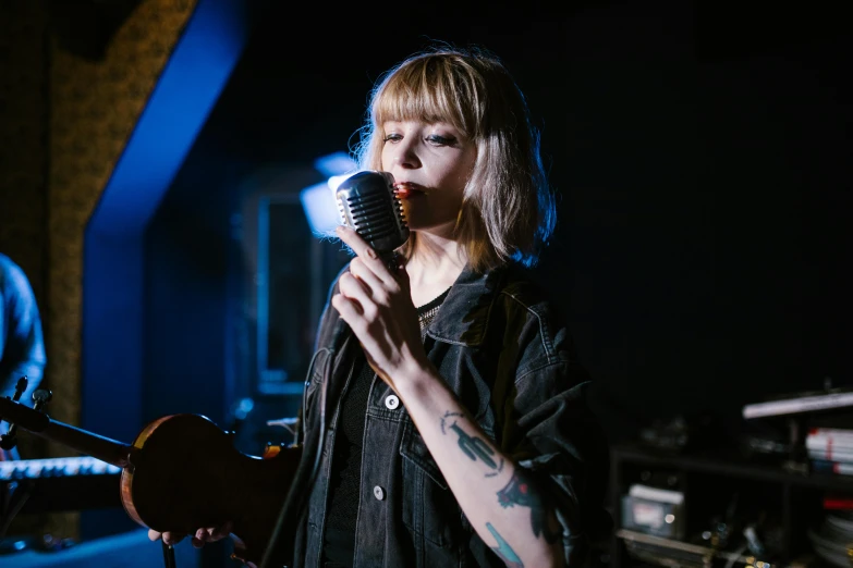a woman singing into a microphone in a dark room, pexels, imogen poots, girl in a record store, holding guitars, eva elfie