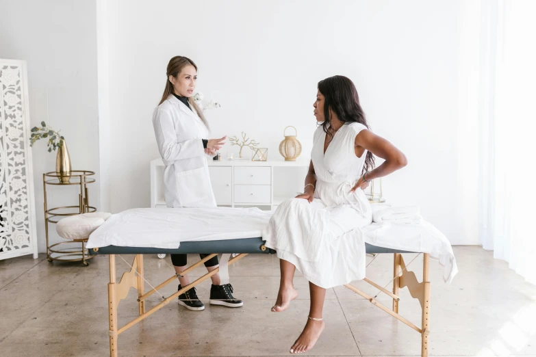 a woman sitting on a massage table next to a woman in a white robe, by Meredith Dillman, trending on unsplash, in front of white back drop, sitting on a lab table, cast, curated collection