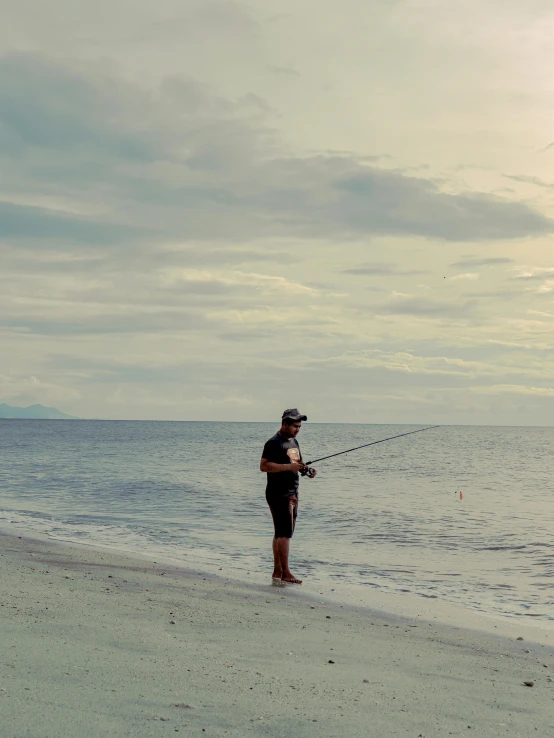 a man standing on top of a beach next to the ocean, fishing pole