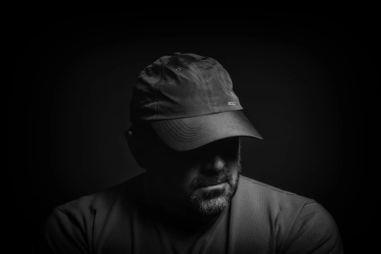 a black and white photo of a man wearing a hat, by Mike Bierek, unsplash, lyco art, dark visor covering top of face, dark. studio lighting, wearing a backwards baseball cap, chris knight