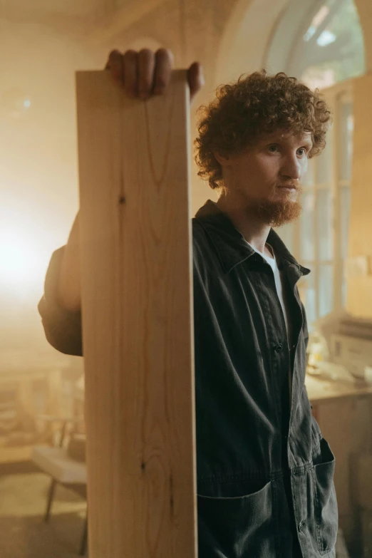 a man standing next to a piece of wood, a portrait, by Jacob Toorenvliet, trending on pexels, renaissance, still from a music video, curly haired, carpenter, light behind
