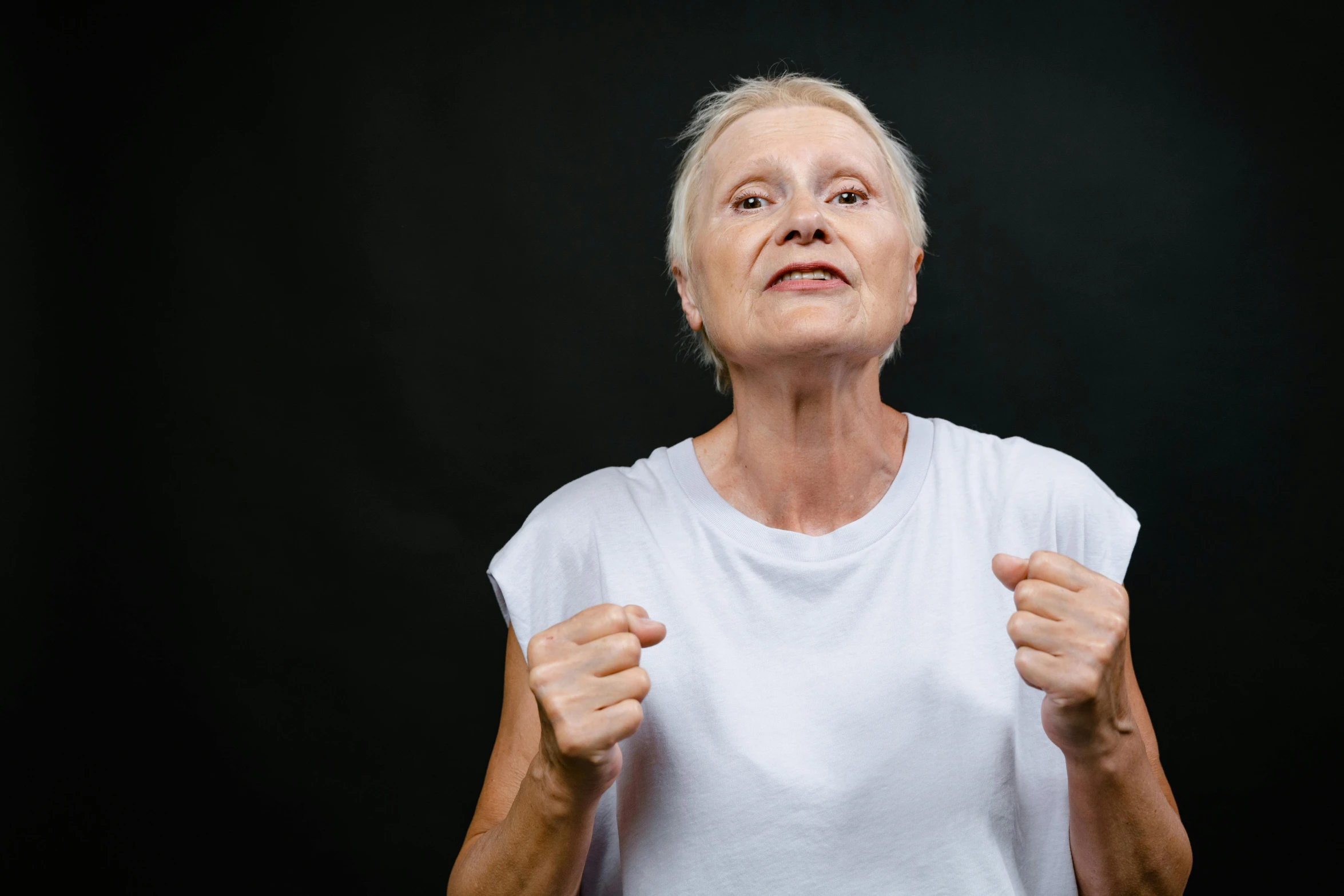 an older woman standing in front of a black background, punching, chest and face, dressed in a white t shirt, dementia