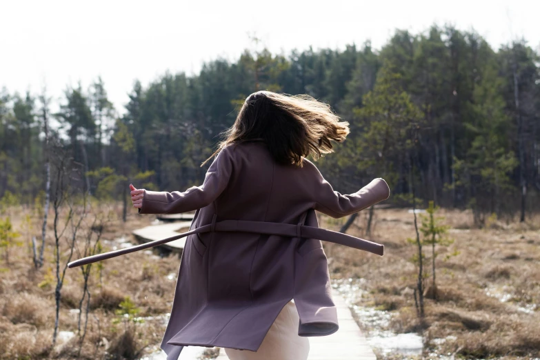 a woman walking across a wooden bridge in a field, by Grytė Pintukaitė, pexels contest winner, wearing a purple detailed coat, whirling, exiting from a wardrobe, long hair windy