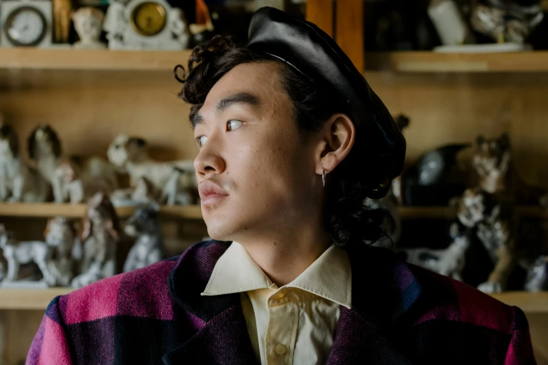 a man standing in front of a shelf filled with figurines, an album cover, by Winona Nelson, trending on pexels, an oversized beret, half asian, androgynous face, curls on top