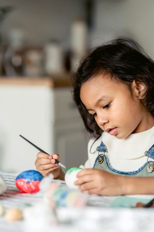 a little girl painting easter eggs on a table, by Elizabeth Durack, pexels contest winner, process art, young asian girl, painting on a badge, little