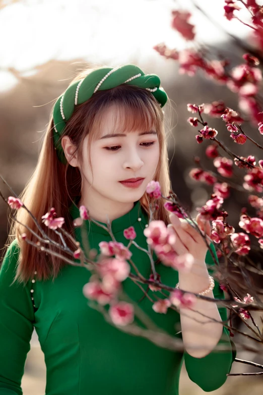 a woman standing in front of a tree with pink flowers, a picture, inspired by Jin Nong, trending on pexels, wearing green clothing, wearing a beret, vietnamese woman, avatar image