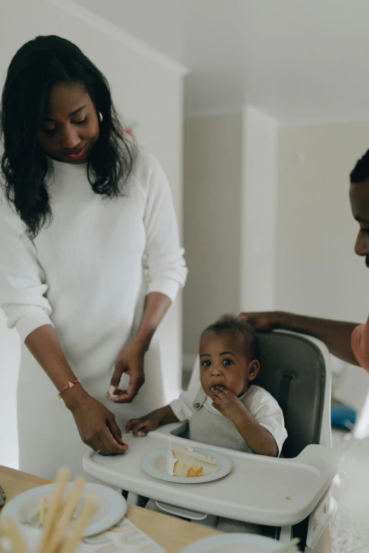 a woman standing next to a baby in a high chair, by Arabella Rankin, pexels contest winner, portrait of family of three, having a snack, white hue, african american