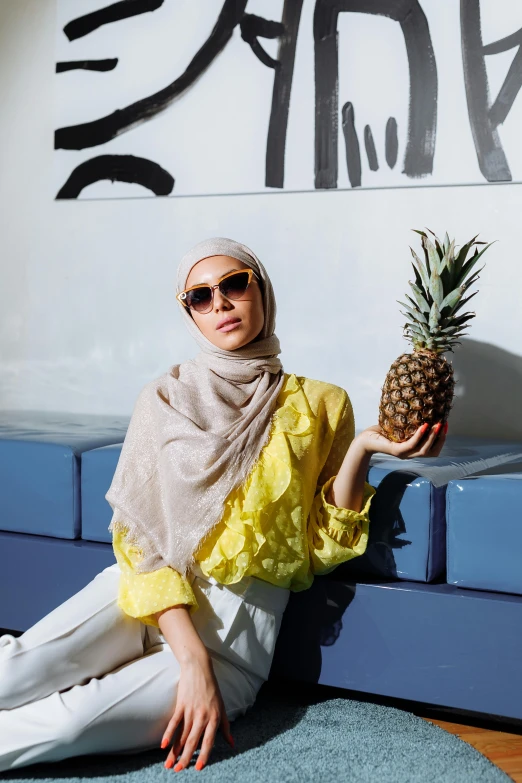 a woman sitting on the floor holding a pineapple, inspired by Charly Amani, hurufiyya, sunglasses and a scarf, light tan, muslim, yellow awning