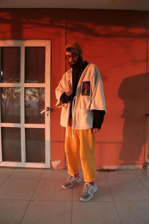 a man standing in front of a red building, an album cover, unsplash, neo-dada, yellow robe, baggy pants, ((sunset)), model posing