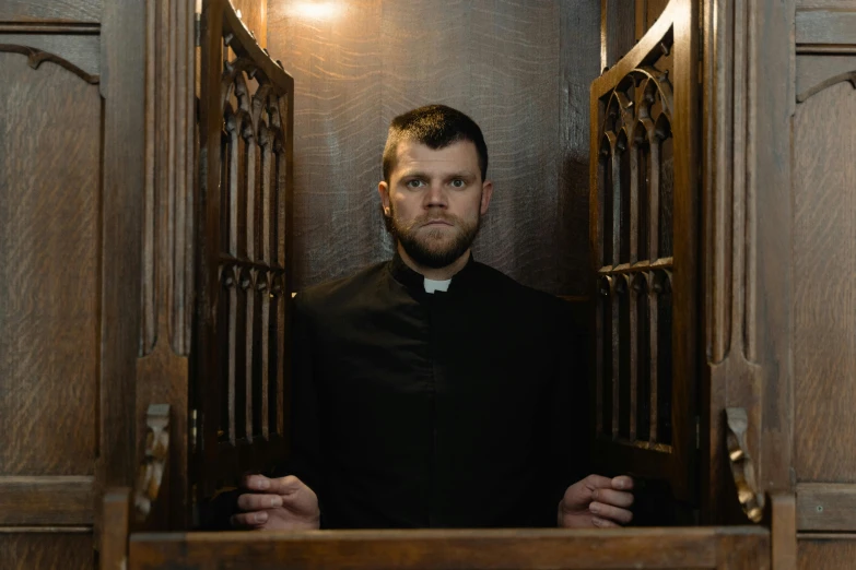 a man that is standing in a doorway, an album cover, by Thomas Fogarty, unsplash, renaissance, priest, seated on wooden chair, intense look, 30 year old man